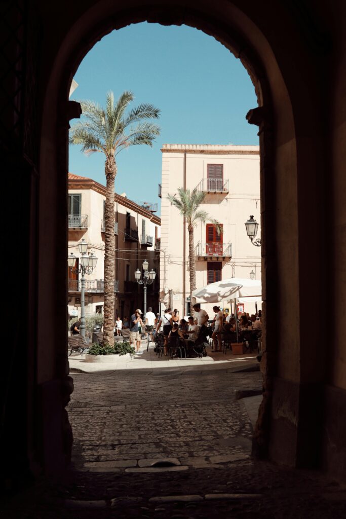 Enjoy the summer vibes through an ancient archway in Cefalù, Sicily, showcasing a vibrant street scene.